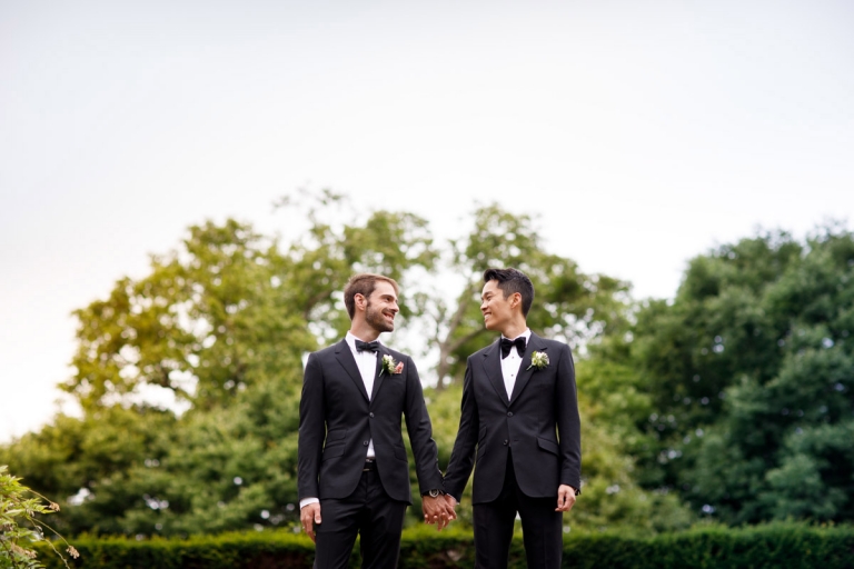 Two grooms look at each other on their wedding day, dressed in black suits. This blog has their English destination wedding at an English castle in the Cotswolds. 