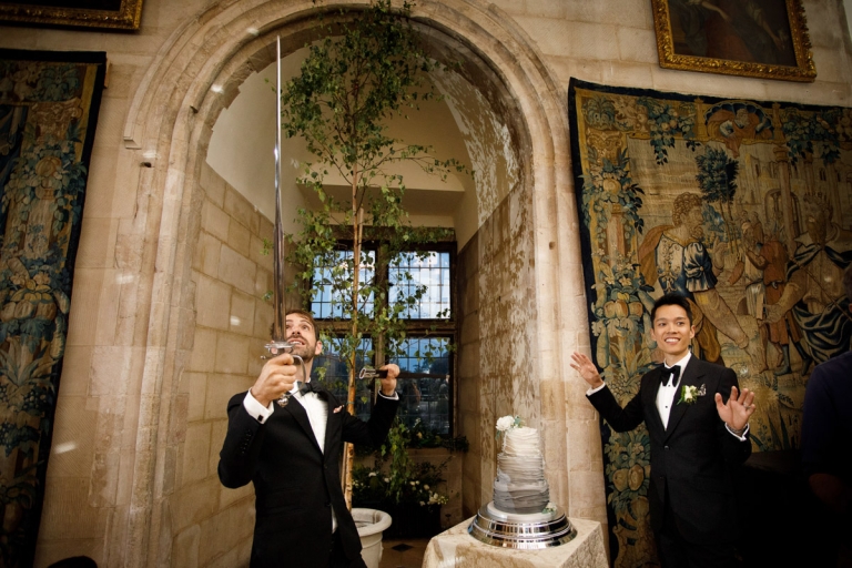 Two grooms about to cut their grey ombre wedding cake with a sword in the archway of the castle surrounded by tapestries. 