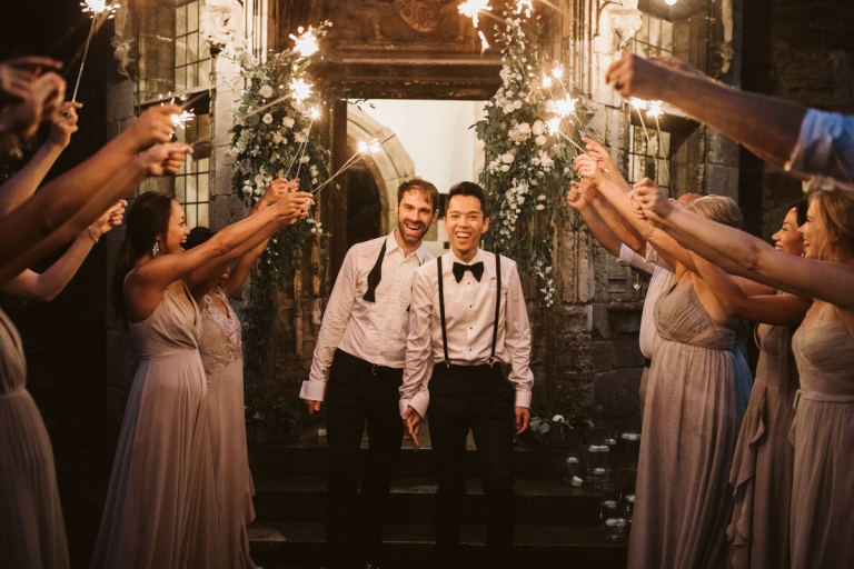 Berkeley Castle Wedding - two grooms smile at camera as their wedding party hold up sparklers