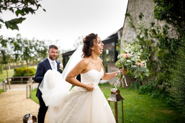 Bride walks back to guests while holding her wild bouquet of roses and green foliage while groom carries the train of her dress