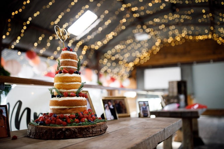 naked wedding Cake decorated with fruit and icing, and a topper with initials, cake table included photos of their family on their own wedding days, with fairy lights above from Cripps Barn stone barn wedding photos