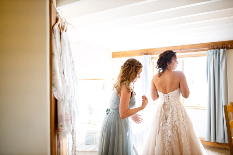 bride hasthe corset of her wedding dress done up by her bridesmaid in blue dress before wedding at cripps barn