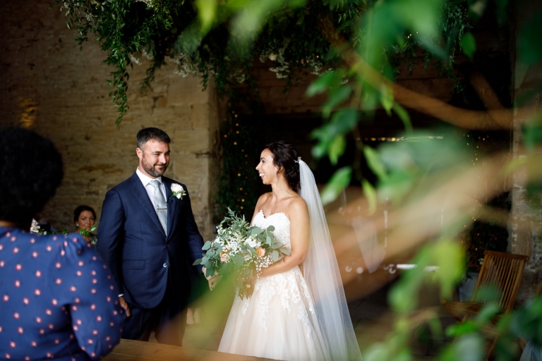 Cripps Stone Barn wedding photos - groom in blue suit and bride carrying her wedding bouquet surrounded by the stone barn and green foliage. 