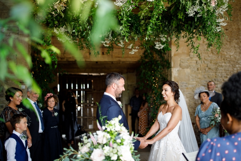 bride and groom hold hands during ceremony at cripps stone barn. They are surrounded by the stone barn and also lots of green foliage and flowers in the foreground and above them on a flower hoop. 