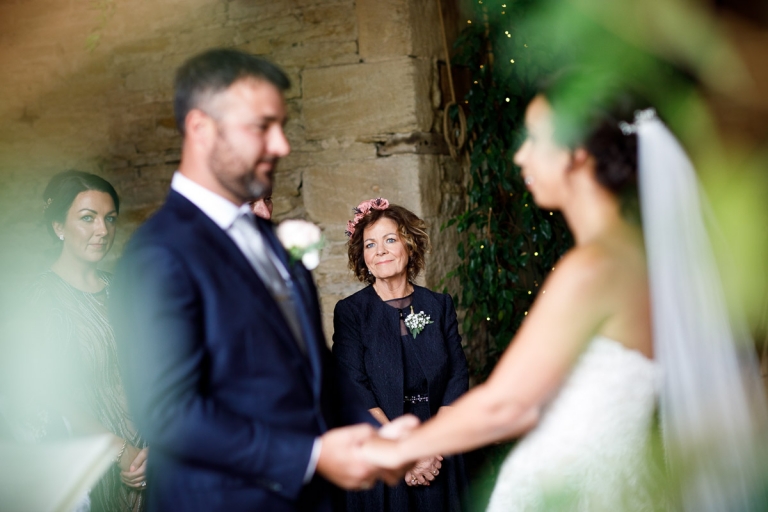 Grooms Mum in dark blue mother of the groom outfit, with pink paper flower crown smiles as she watches her Son getting married at Stone Barn
