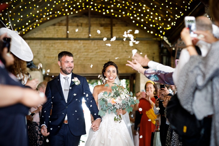 Couple walk through guests throwing confetti with fairy lights above at Cripps Stone Barn