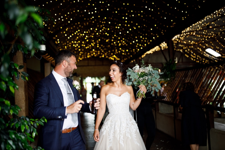 Couple walk through fairy lights at Cripps stone Barn, bride holding her bouquet with roses and lots of freen foliage which creates a wild and rustic bouquet. 
