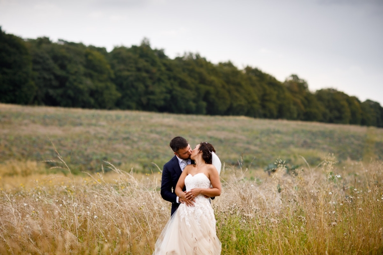 Couple kiss in English landscape with lots of lush trees and long grass at Stone Barn