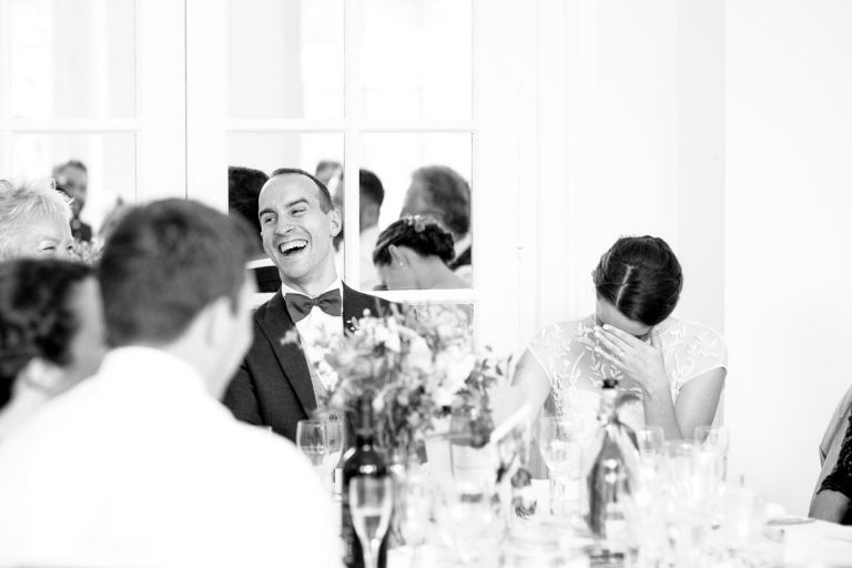 bride shields her face as the groom laughs at a joke from the bestman at their wedding