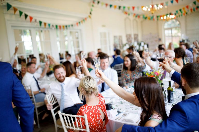 inside the orangery at goldney hall, all guests raise their glasses to toast the bride and groom, lots of colourful bunting in the background