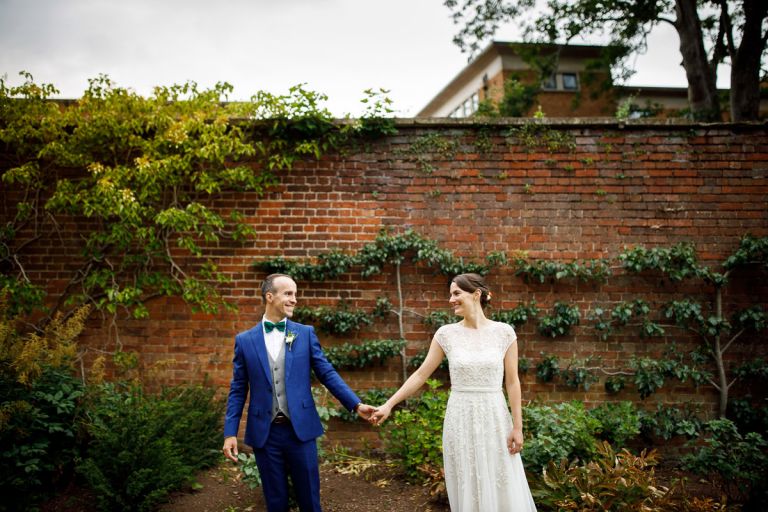 Couple stand hand in hand and smile at each other in front of red brick wall with trees growing up it in the garden of goldney house