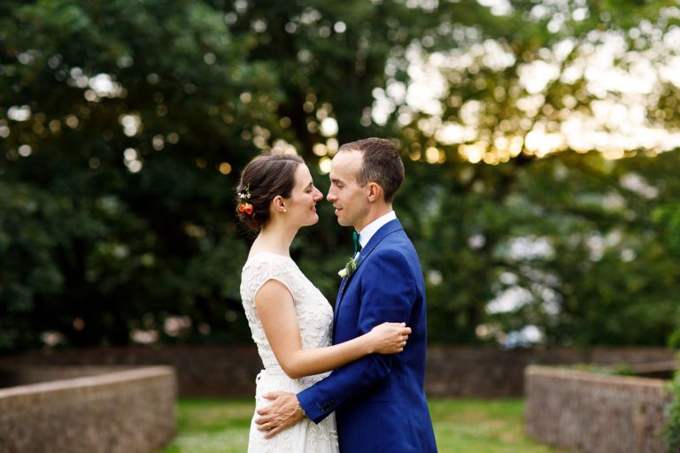 Couple stand very close and look into each others eyes with trees behind them, sun is setting behind the trees which gives an orange glow at goldney hall