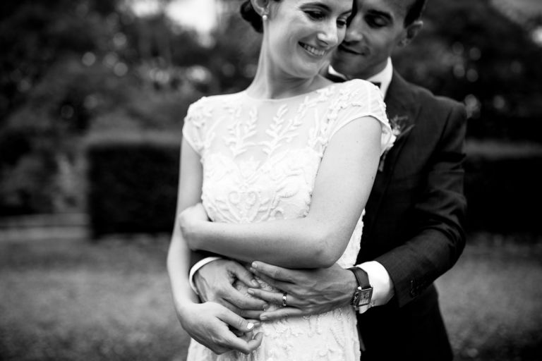 close up photo of groom hugging bride from behind, he nuzzles into her and he holds his wedding ring finger as he does