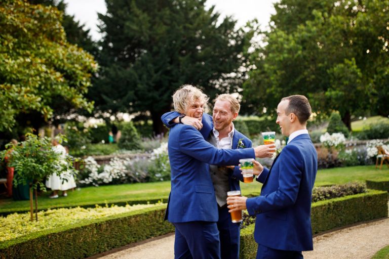 groom and groomsmen mess around laughing with drinks in the gardens at goldney hall, they wear matching blue suits