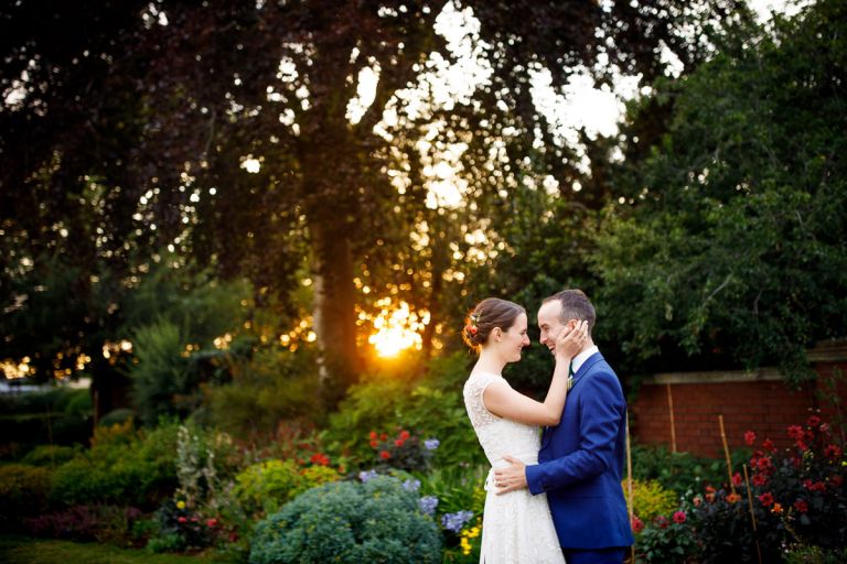 bride holds grooms head in her hands as they laugh, the low evening sun is golden and coming through the trees in the background at a garden wedding in bristol