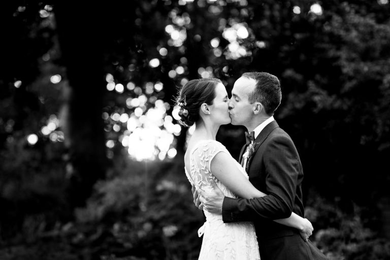 Couple embrace and kiss with the ligh sparkling through the trees behind in black and white in the city garden of goldney house
