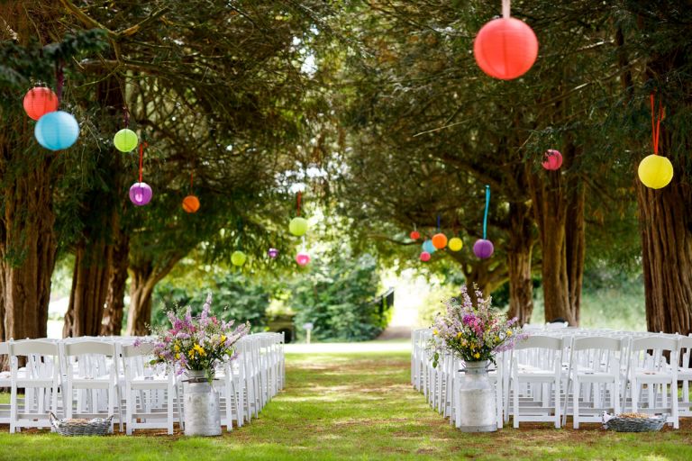Outside wedding at goldney hall, chairs set up under the trees where colourful lanterns hang and flowers sit at the edge of the aisles in milk churns