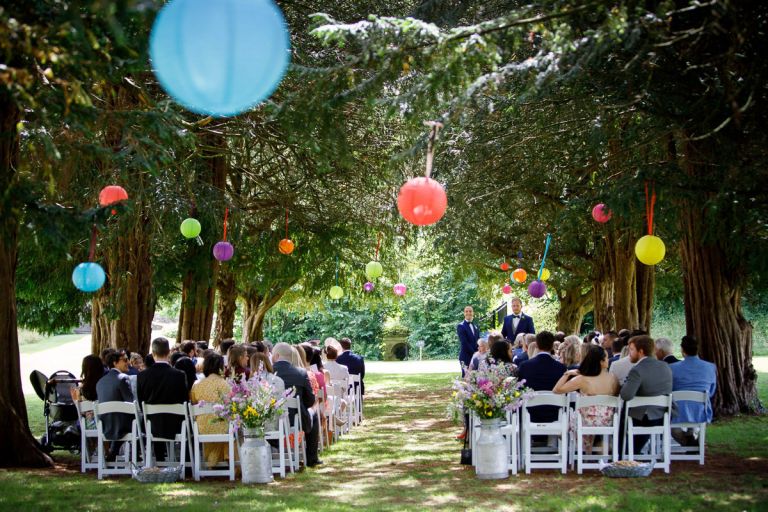 view of guests sat waiting under the trees for the outside wedding at goldney hall, groom and bestman in distance waiting for the bride