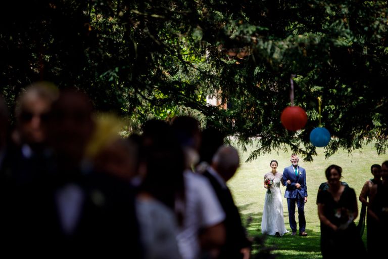 bride is escorted into her outside wedding at goldney hall by her dad, she is in bright sunlight while everyone else is in the shade