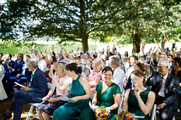 lots of guests hold up cards and laugh during the mr and mrs game taking place during the outdoor wedding ceremony in the trees in bristol