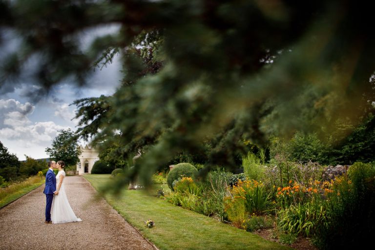 Couple kiss in the pathway surrounded by flower beds and lots of tree branches in the foreground in the gardens in bristol wedding