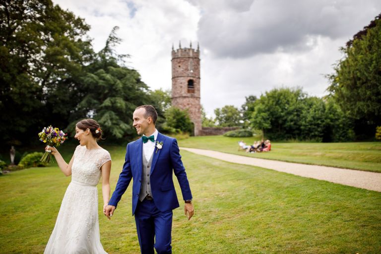 Couple wave and smile at guests as they walk back to their wedding at goldney hall with the tower in the background