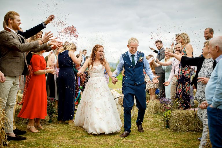 Couple walk back up the aisle laughing as a lot of confetti is thrown at them after their outside humanist wedding