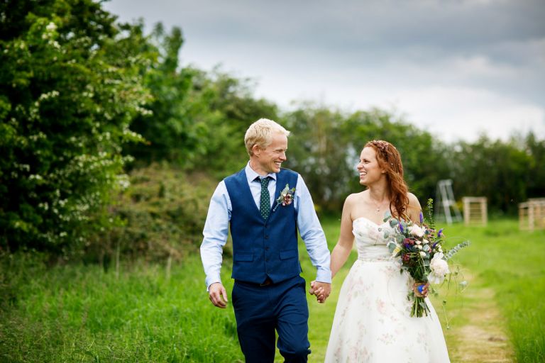 Couple walk hand in hand smiling at each other after their ceremony, bride is wearing a dress with flowers on while holding a wild bouquet of flowers