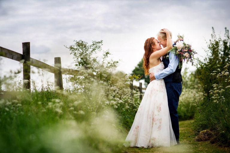 Bride and groom kiss in wild flowers, the bride brings her hand up to the grooms head to hug him as they go to kiss. 