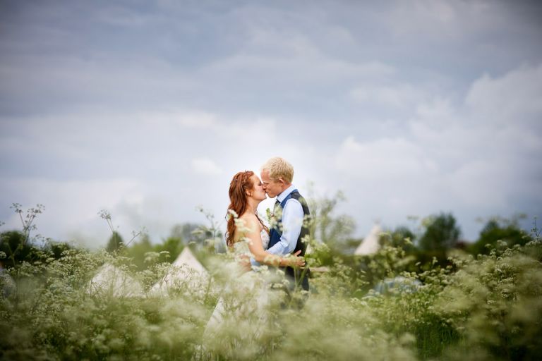 Couple kiss in the wild flowers with their tipis and tents in the background during their humanist outside wedding