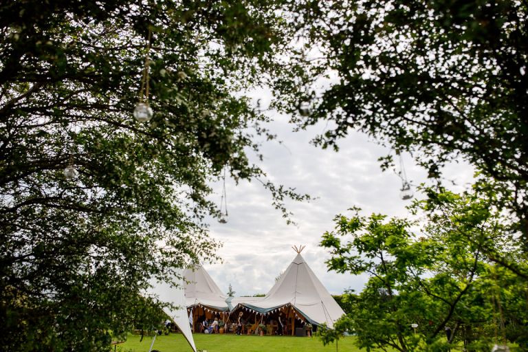 Wedding tipi full of guests from a distance framed by trees with decorations hanging in them