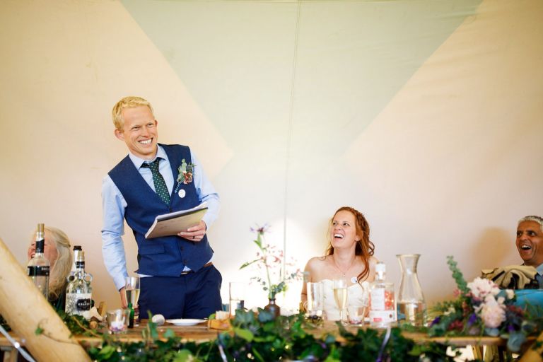 Groom is standing to give his speech, bride and her dad is laughing