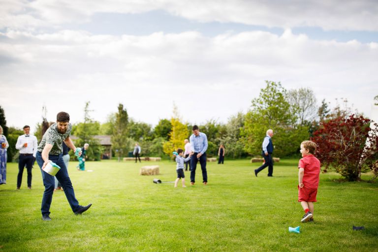 Kid and his dad play a game during the outside wedding reception, he tosses a ball that has to be caught in a can. 