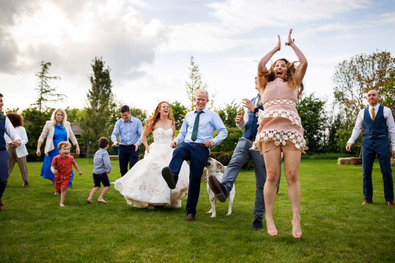 Enthusiastic guest jumps in the air to clap during ceilidh while wedding couple laugh in the background