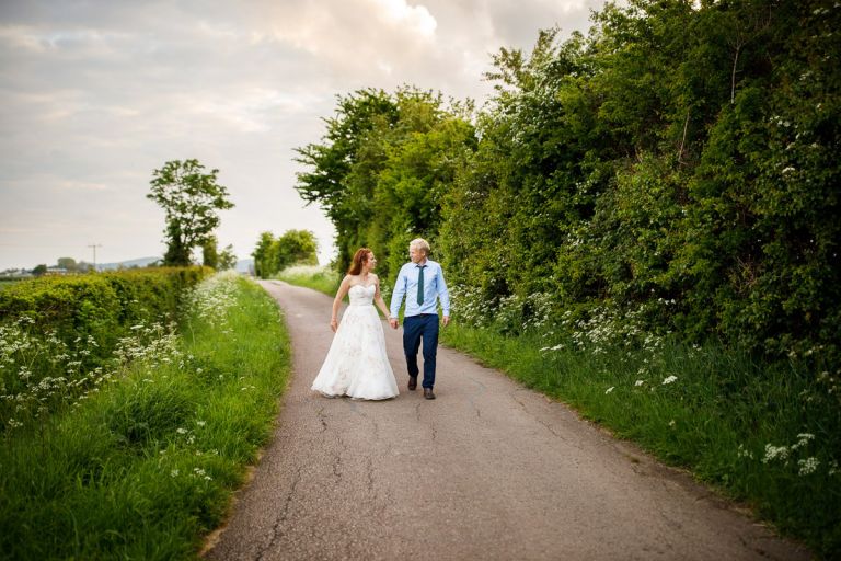 Wedding couple wander down pretty countryside lane holding hands