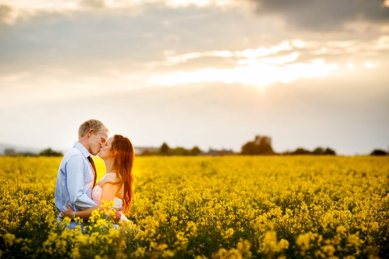 Couple kiss in beautiful yellow field of flowers while the sun gets low in the sky behind them given a lens flare