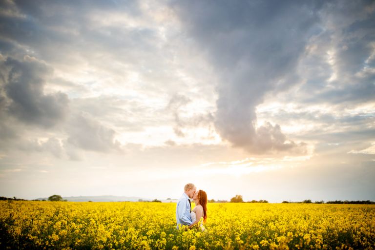 Couple surrounded by a field of yellow flowers, the sky is dramatic with lots of clouds and some golden light as the sun gets low