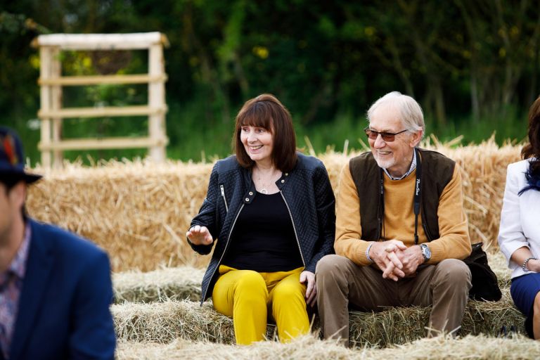 Wedding guest sit on haybales waiting for a wedding ceremony to start, they are laughing. 