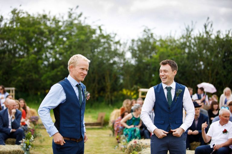 Groom smiles as his bride arrives in the distance while his bestman laughs next to him. 