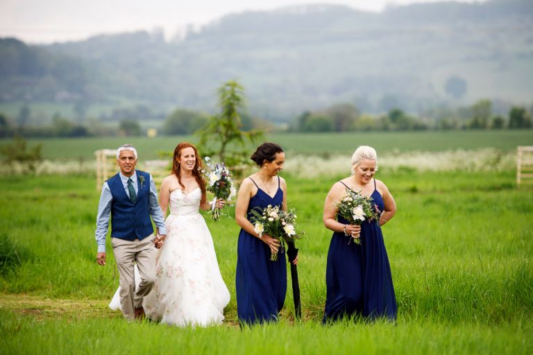 Bride arrives and walks through the field to the ceremony with her dad and her two bridesmaids. 