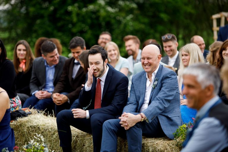 Guests laugh during humanist ceremony while sat on haybales at the outdoor wedding ceremony