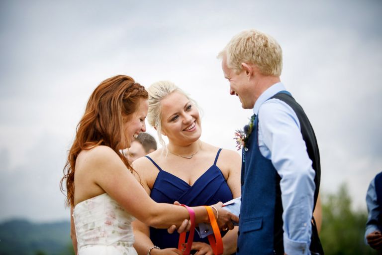 Bridesmaid ties ribbon around the arms of bride and groom during handfasting. 