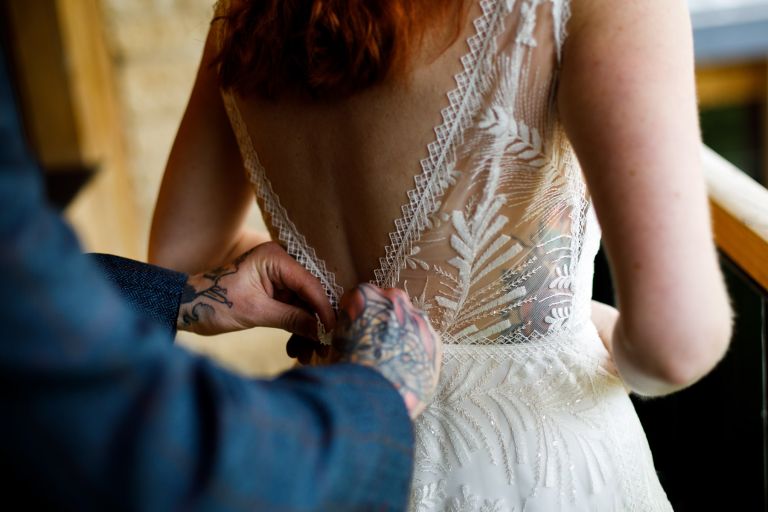 Detail of brides dress as her groom does it up, can see dress detail, grooms jacket and tattoos on both people