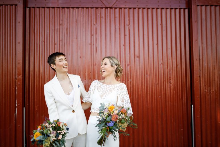 Brides laugh together in front of red M Shed doors at same sex Bristol wedding flowers from Ivory Flowers