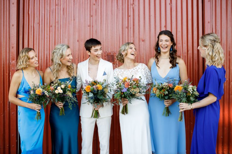 Brides laugh with their wedding party in front of red metal doors
